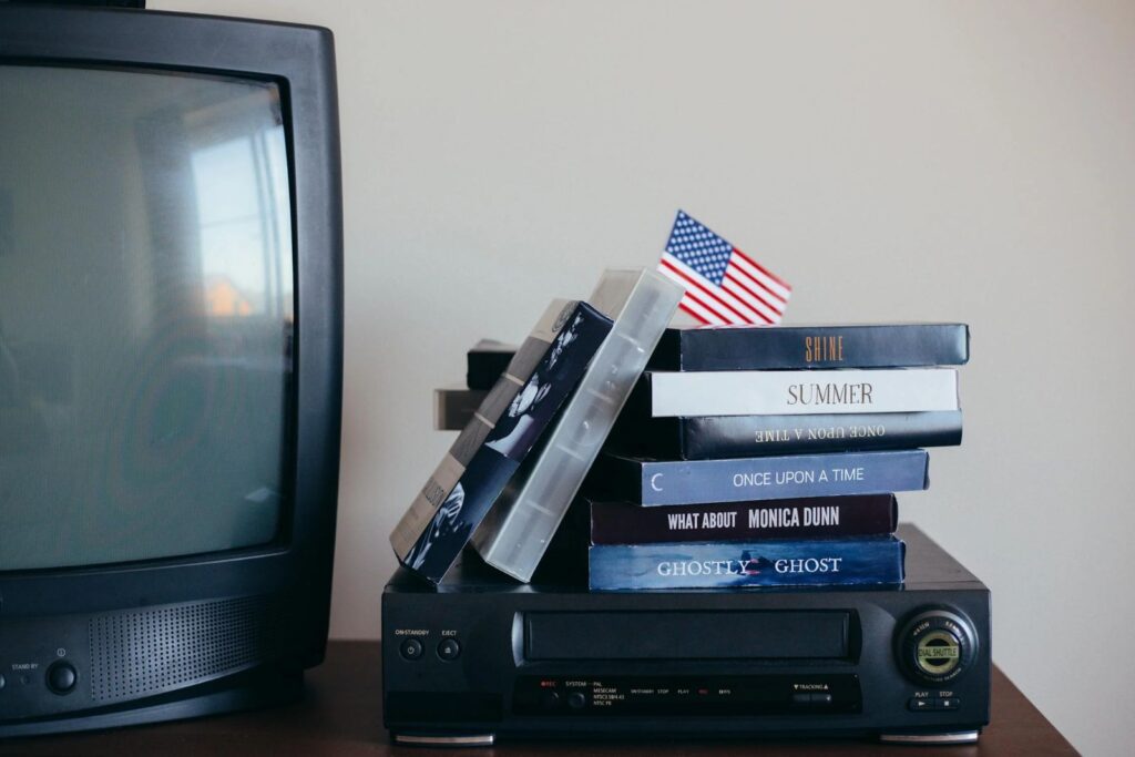 Junto a un televisor y un reproductor de vídeo antiguos sobre una mesa de madera hay una pila de cintas VHS que evocan nostalgia en el acogedor hogar Entre las cintas hay una pequeña bandera estadounidense cuidadosamente colocada-Lo que no debes tener en tu casa despues de los 30: es hora de transformar tu hogar