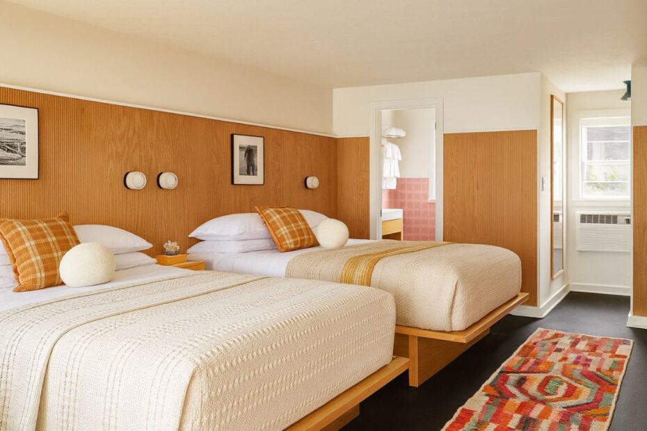 Una habitación de hotel con dos camas dobles paredes con paneles de madera fotos enmarcadas una alfombra de colores y una puerta abierta al cuarto de baño: perfecta para quienes se inspiran en el diseño de interiores de Airbnb-Diseño de Airbnb: consejos prácticos para destacar tu propiedad