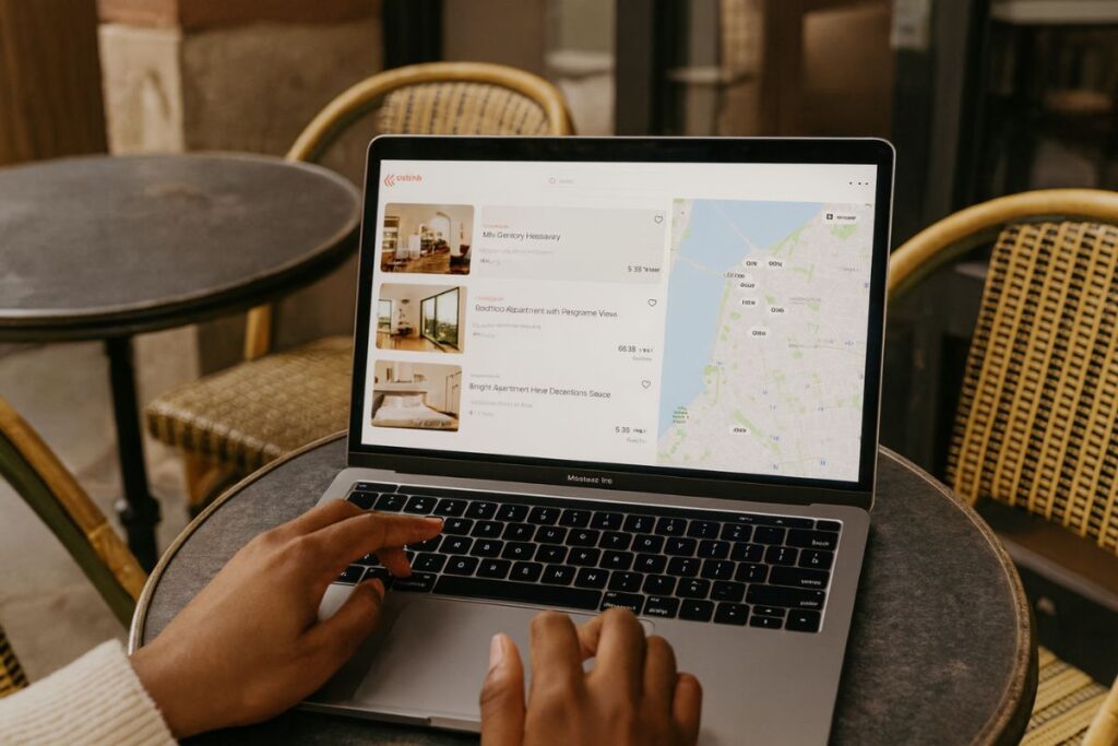 Una persona utiliza un ordenador portátil en la mesa de un café al aire libre navegando por los listados de alojamientos con un mapa desplegado en la parte derecha de la pantalla y buscando consejos prácticos sobre diseño de Airbnb. diseño airbnb