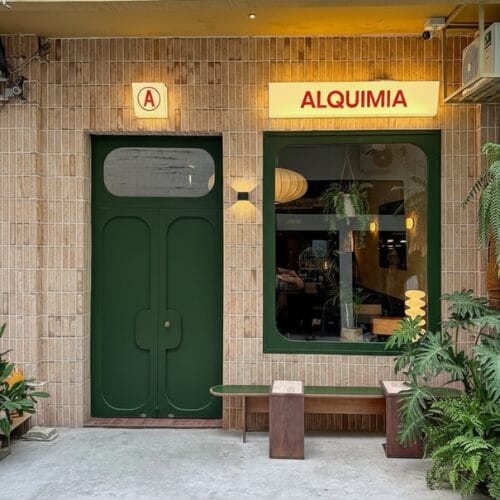 El exterior de la cafetería "Alquimia" destaca por su arquitectura con una puerta verde gran ventanal banco plantas en macetas y un letrero iluminado sobre la fachada de ladrillo beige-Diseño de Interiores Online