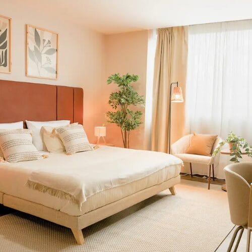 Una cama bien hecha con ropa de cama beige en un dormitorio luminoso muestra un interior de diseño elegante con una planta en maceta una silla junto a la ventana y arte botánico enmarcado en la pared-Diseño de Interiores Online